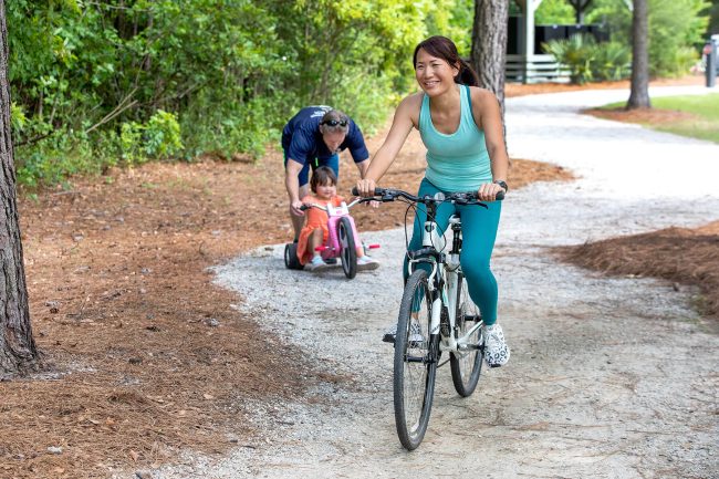 A father pushes his daughter on three wheeler while mom bikes ahead on a gravel trail